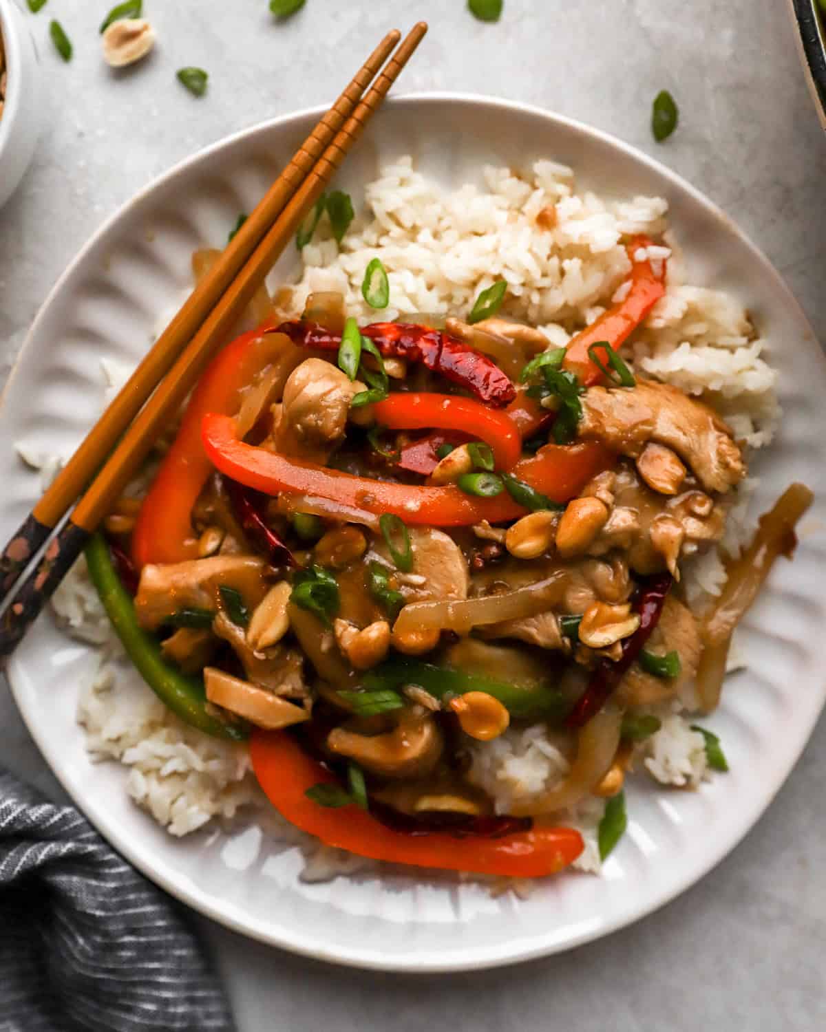 A plate of Szechuan chicken served over steamed white rice, garnished with green onions and accompanied by wooden chopsticks.