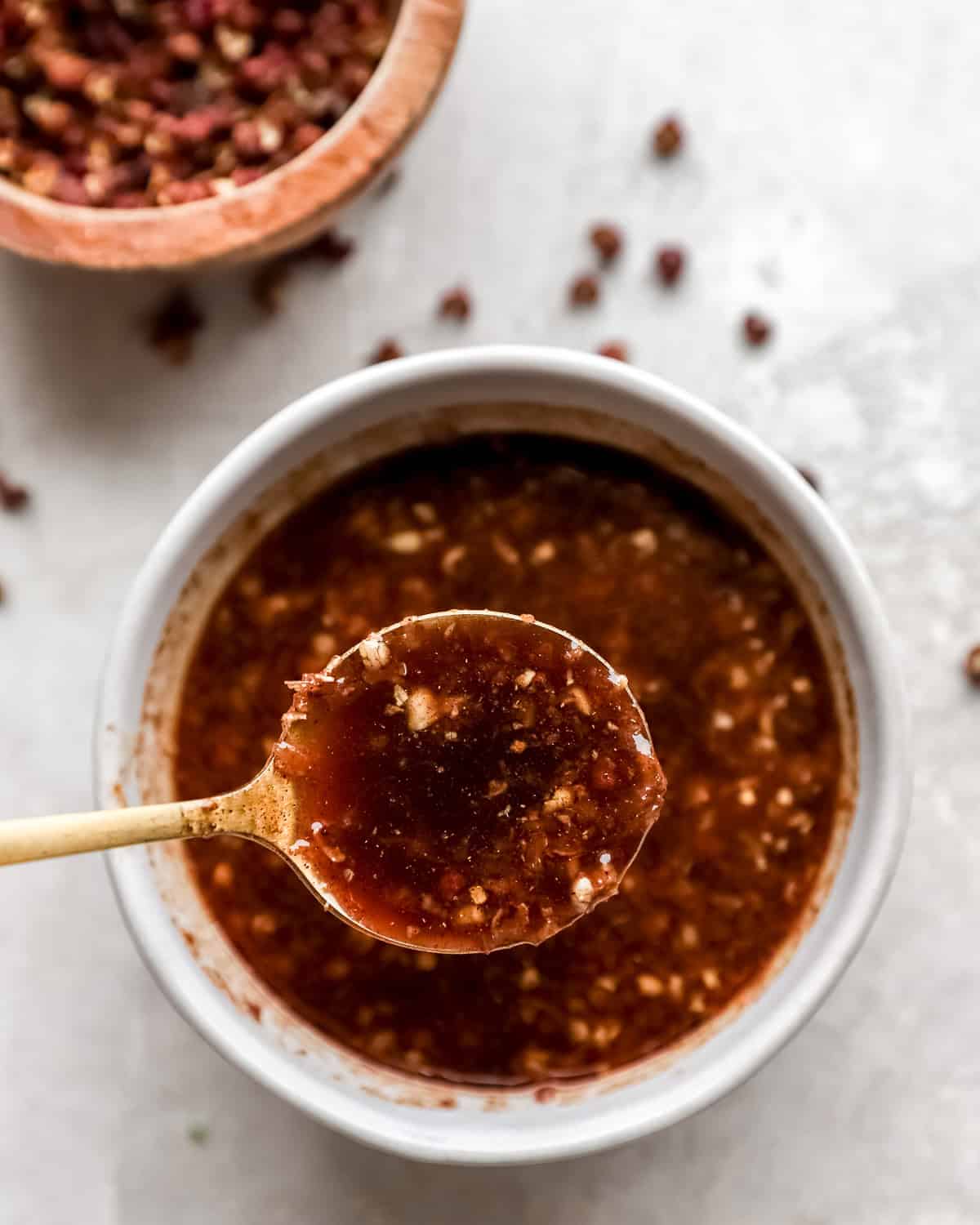 A small white bowl of rich, homemade Szechuan sauce, topped with minced ingredients, with a spoon lifting some sauce for a close-up