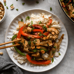 Top view of a plate of Szechuan chicken with colorful vegetables and peanuts, served on a bed of rice, surrounded by small bowls of garnishes.