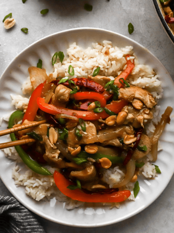 Top view of a plate of Szechuan chicken with colorful vegetables and peanuts, served on a bed of rice, surrounded by small bowls of garnishes.