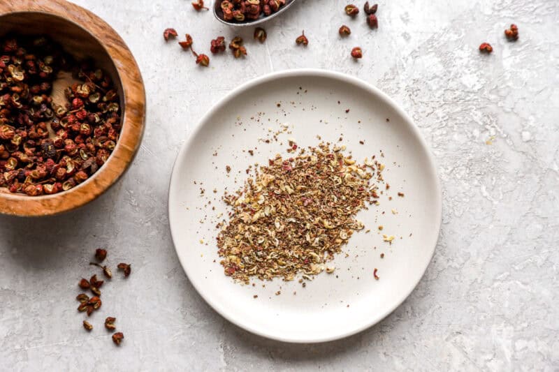A close-up of ground Szechuan peppercorns on a white plate, with whole peppercorns and a wooden bowl in the background.