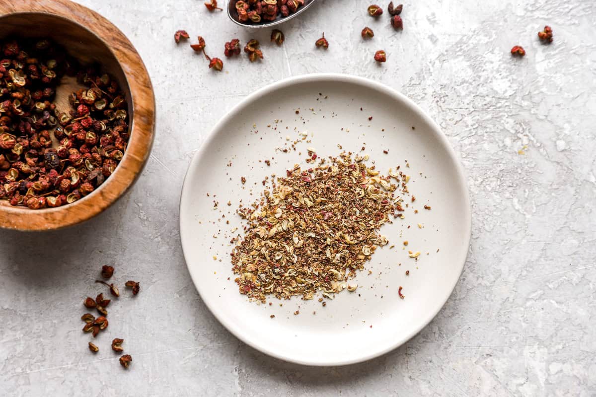 A close-up of ground Szechuan peppercorns on a white plate, with whole peppercorns and a wooden bowl in the background.