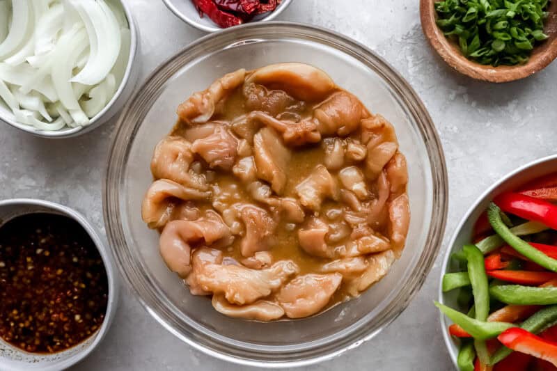 Raw chicken marinating in a bowl surrounded by bowls of sliced onions, bell peppers, dried chili peppers, and sauce ingredients.