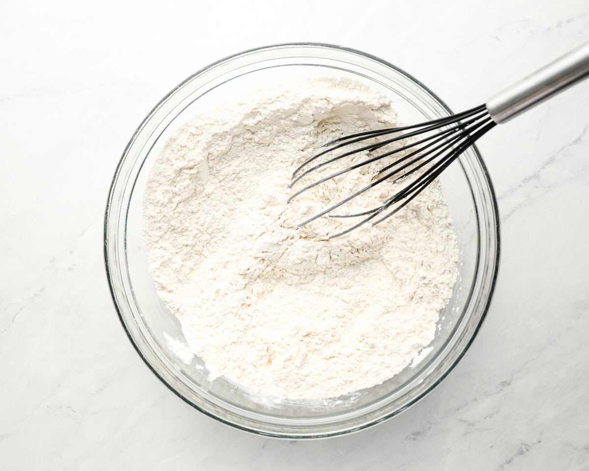 A glass bowl filled with flour and a whisk, showing the beginning stage of the sopapilla dough preparation.