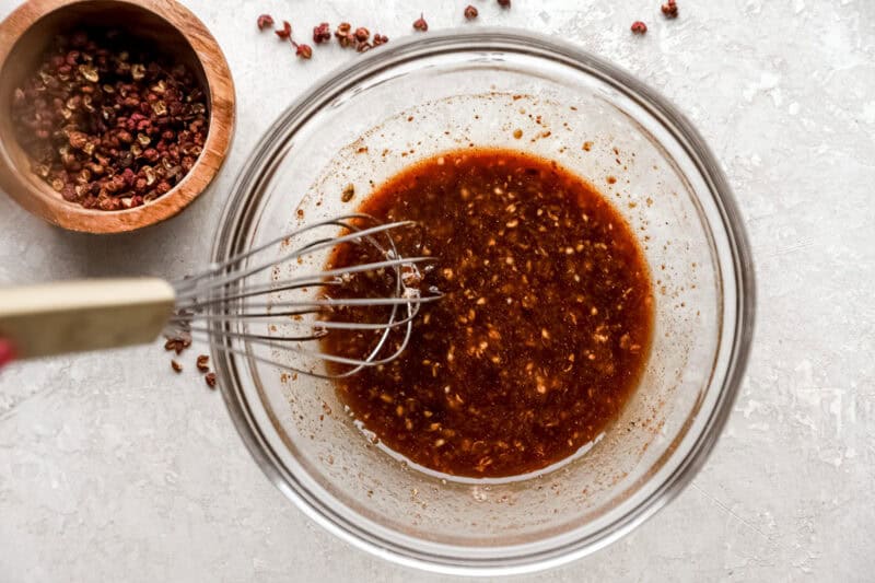 A glass bowl filled with freshly whisked Szechuan sauce, surrounded by whole peppercorns and a wooden bowl of spices on a textured light surface