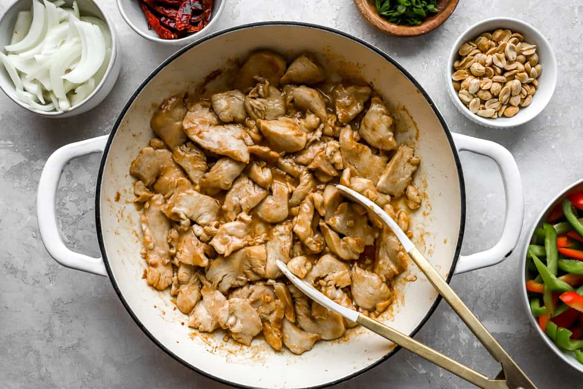 Cooked chicken pieces in a white skillet, surrounded by bowls of sliced vegetables, dried chili peppers, peanuts, and green onions.
