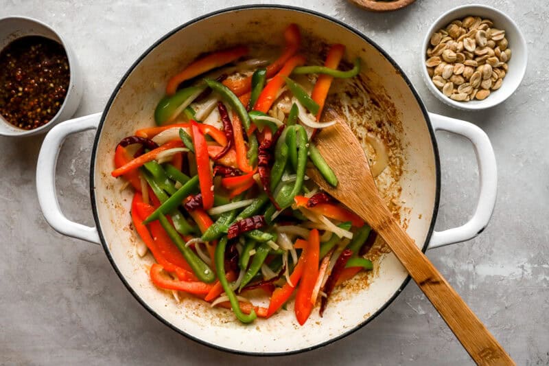 Bell peppers and onions being stir-fried in a skillet with dried chili peppers, surrounded by bowls of peanuts and sauce.
