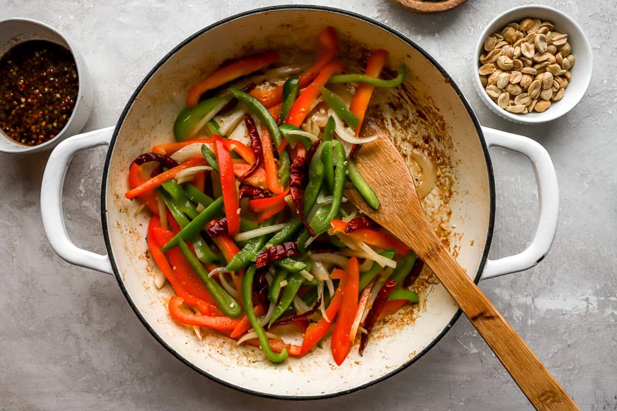 Bell peppers and onions being stir-fried in a skillet with dried chili peppers, surrounded by bowls of peanuts and sauce.