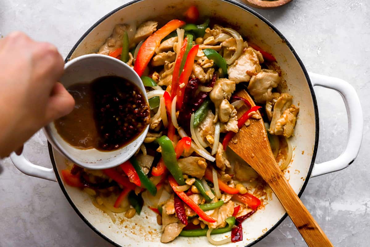 Szechuan sauce being poured into a skillet filled with cooked chicken, vegetables, and peanuts, highlighting the final cooking step.