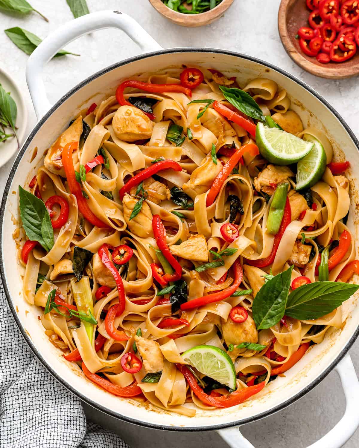 Overhead view of a skillet filled with cooked drunken noodles, chicken, red bell peppers, Thai basil, and lime wedges.