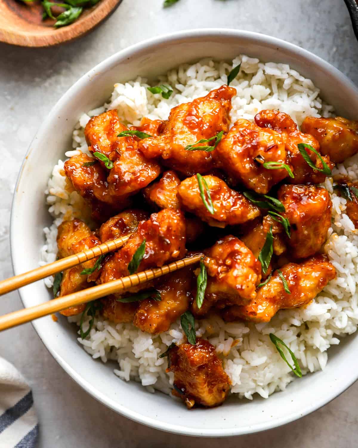 Close-up shot of sweet and spicy General Tso’s Chicken on a bed of rice, topped with green onions and served with chopsticks.
