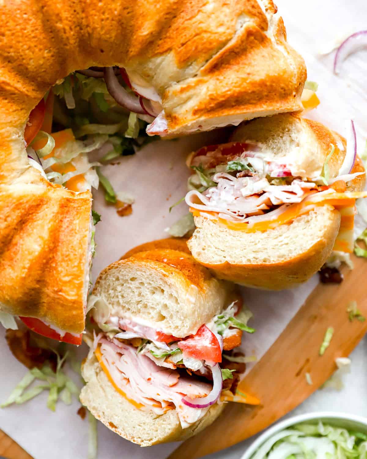 A freshly baked Bundt-shaped sandwich loaf is sliced open, revealing layers of deli meats, cheese, lettuce, tomatoes, and mayonnaise. The sandwich is cut into pieces, with fresh ingredients spilling onto the cutting board.
