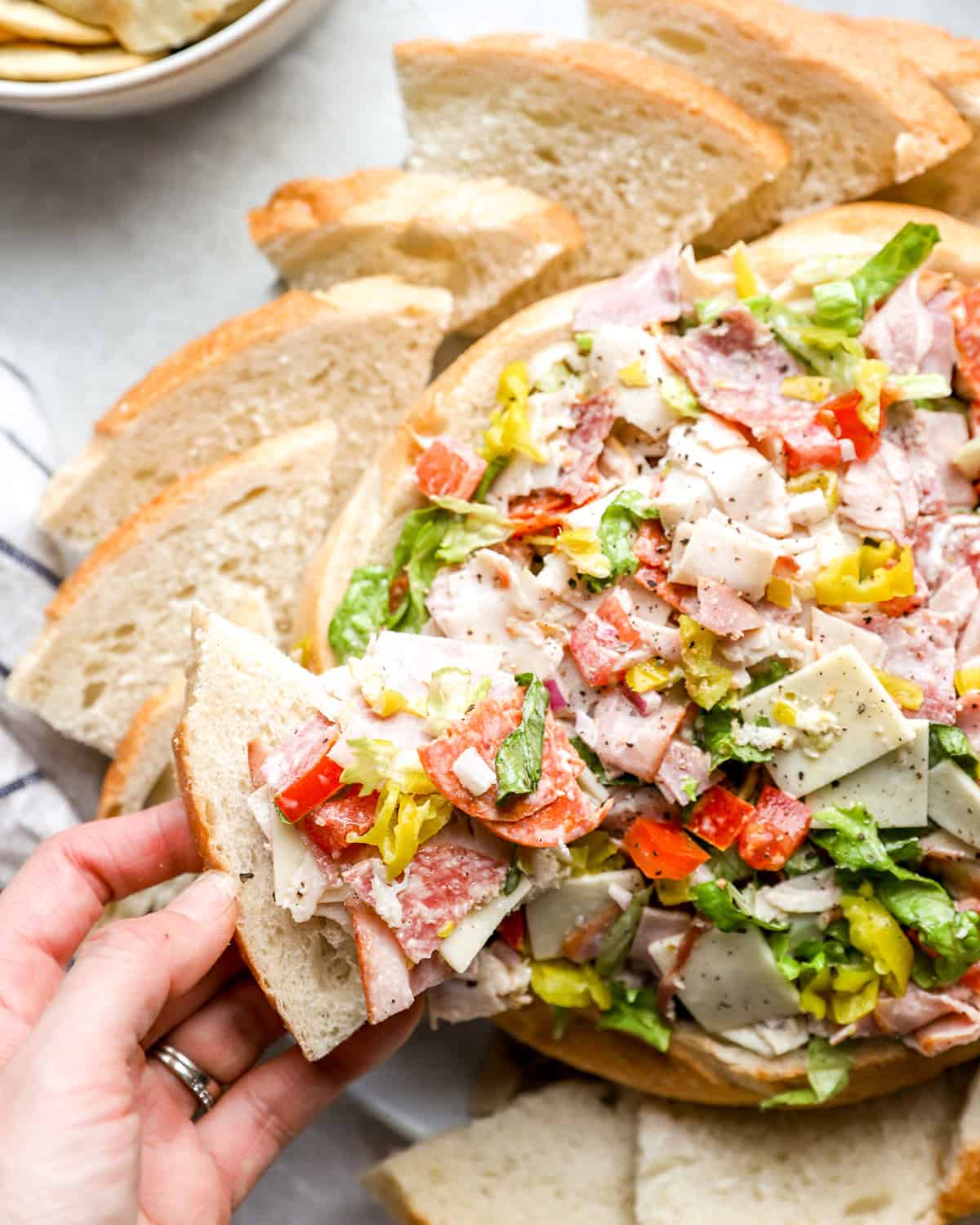 A hand holding a slice of crusty Italian bread topped with hoagie dip, featuring layers of deli meats, provolone cheese, lettuce, tomatoes, and pepperoncini.