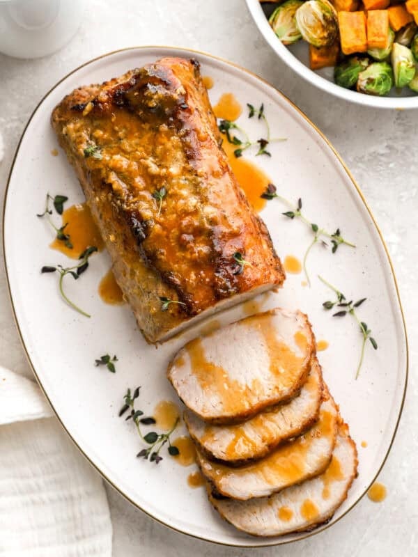 A beautifully roasted pork loin on a white oval platter, sliced and drizzled with a golden maple Dijon glaze, garnished with fresh thyme. A side dish of roasted Brussels sprouts and sweet potatoes is partially visible in the background.