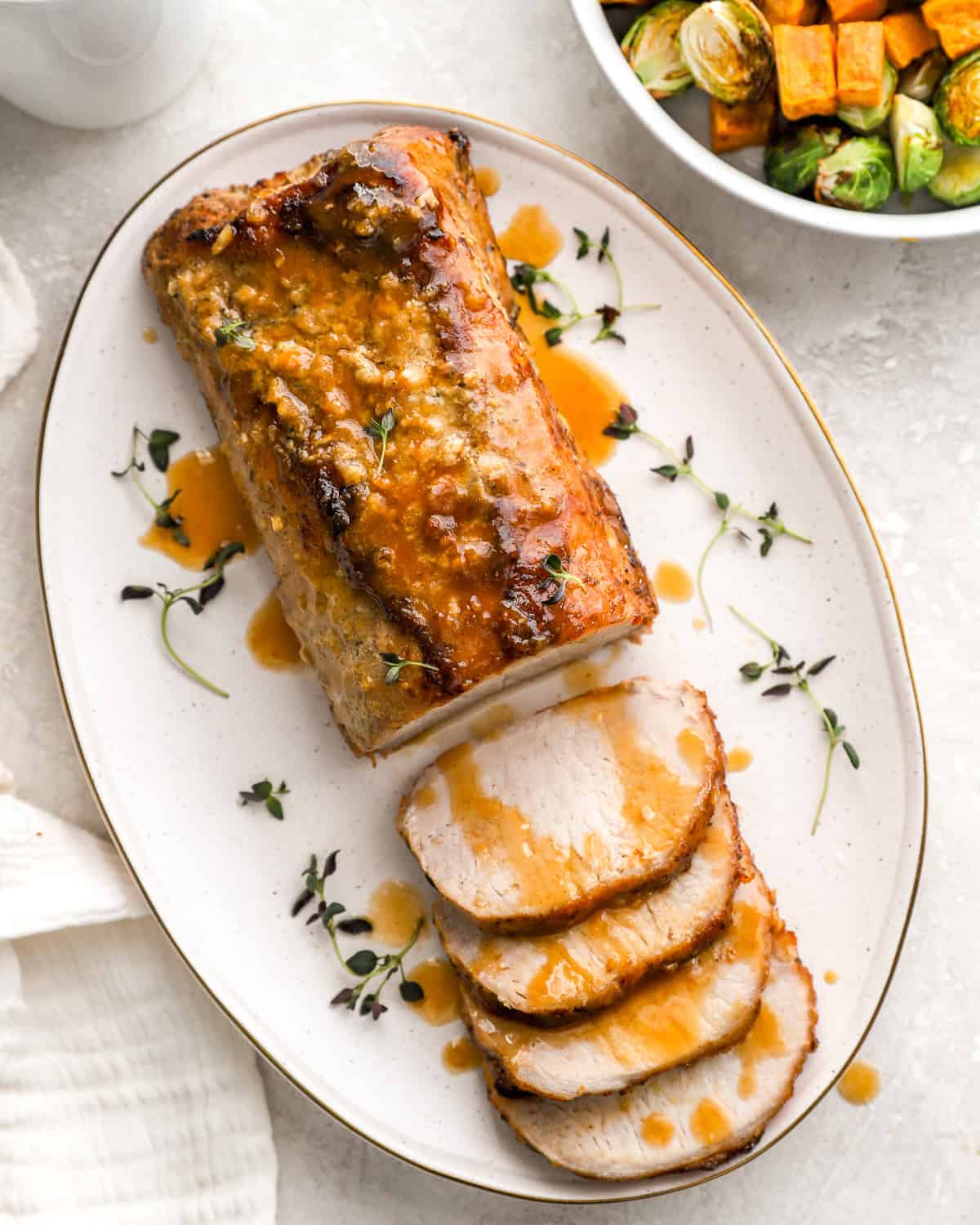A beautifully roasted pork loin on a white oval platter, sliced and drizzled with a golden maple Dijon glaze, garnished with fresh thyme. A side dish of roasted Brussels sprouts and sweet potatoes is partially visible in the background.