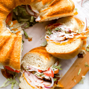 A close-up of the finished party sandwich, cut into sections, highlighting the crispy bacon, melted cheese, fresh vegetables, and soft bread layers.