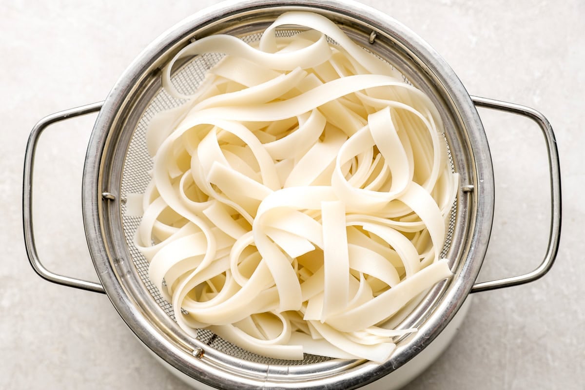 Cooked wide rice noodles in a metal colander, freshly drained and ready for stir-frying.