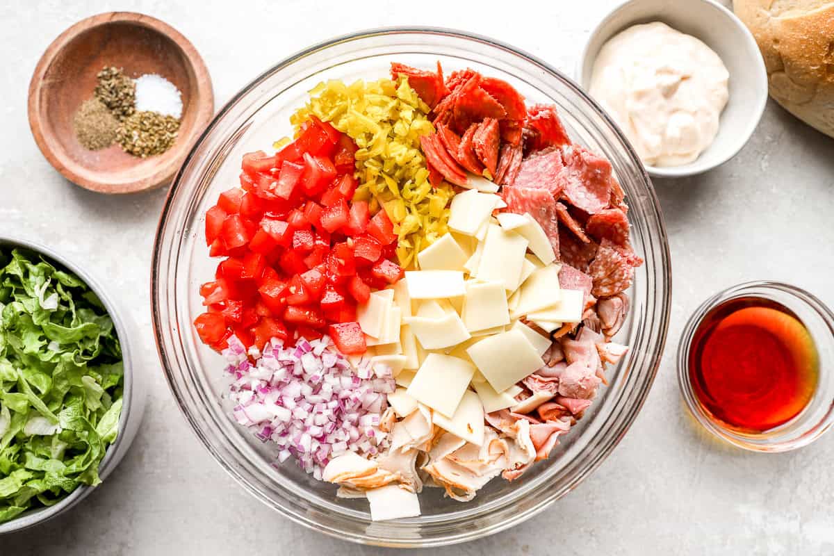 A glass mixing bowl filled with chopped deli meats, provolone cheese, diced tomatoes, chopped red onion, and pepperoncini, ready to be mixed.