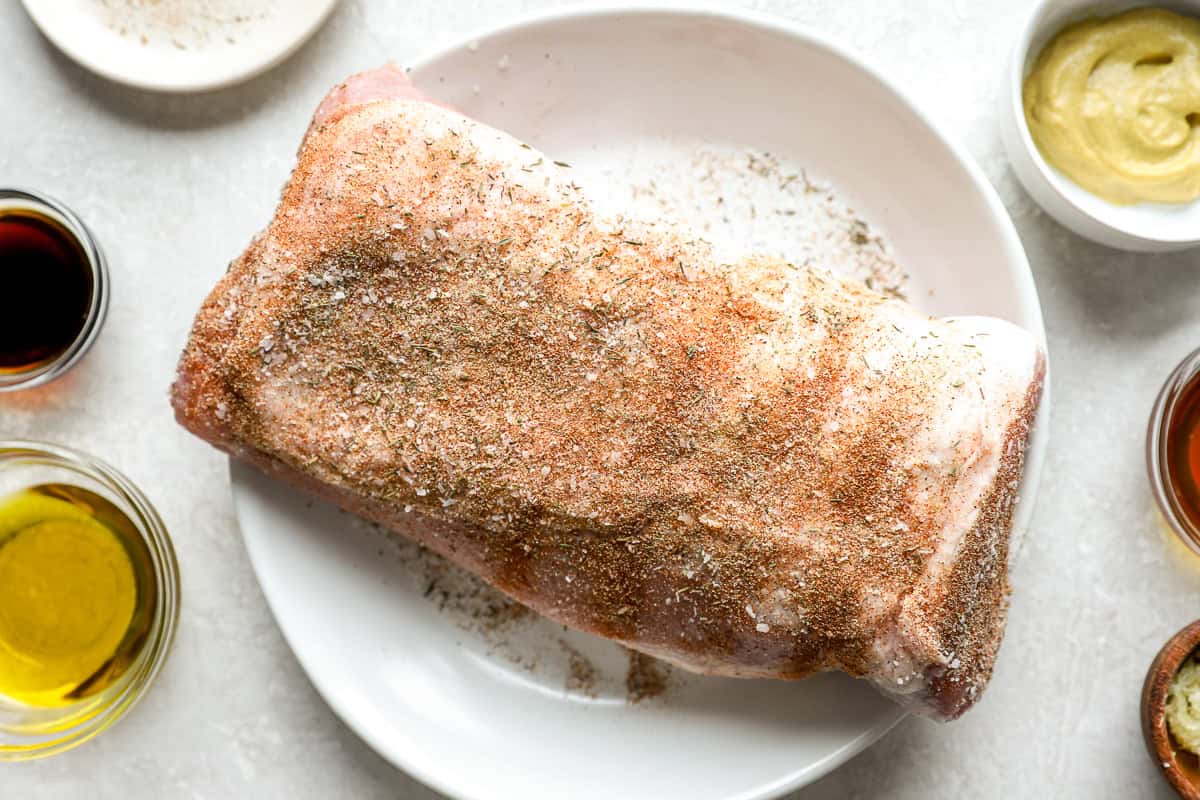 A raw pork loin coated in a dry rub seasoning mix on a white plate, surrounded by small bowls of maple syrup, Dijon mustard, soy sauce, and olive oil.