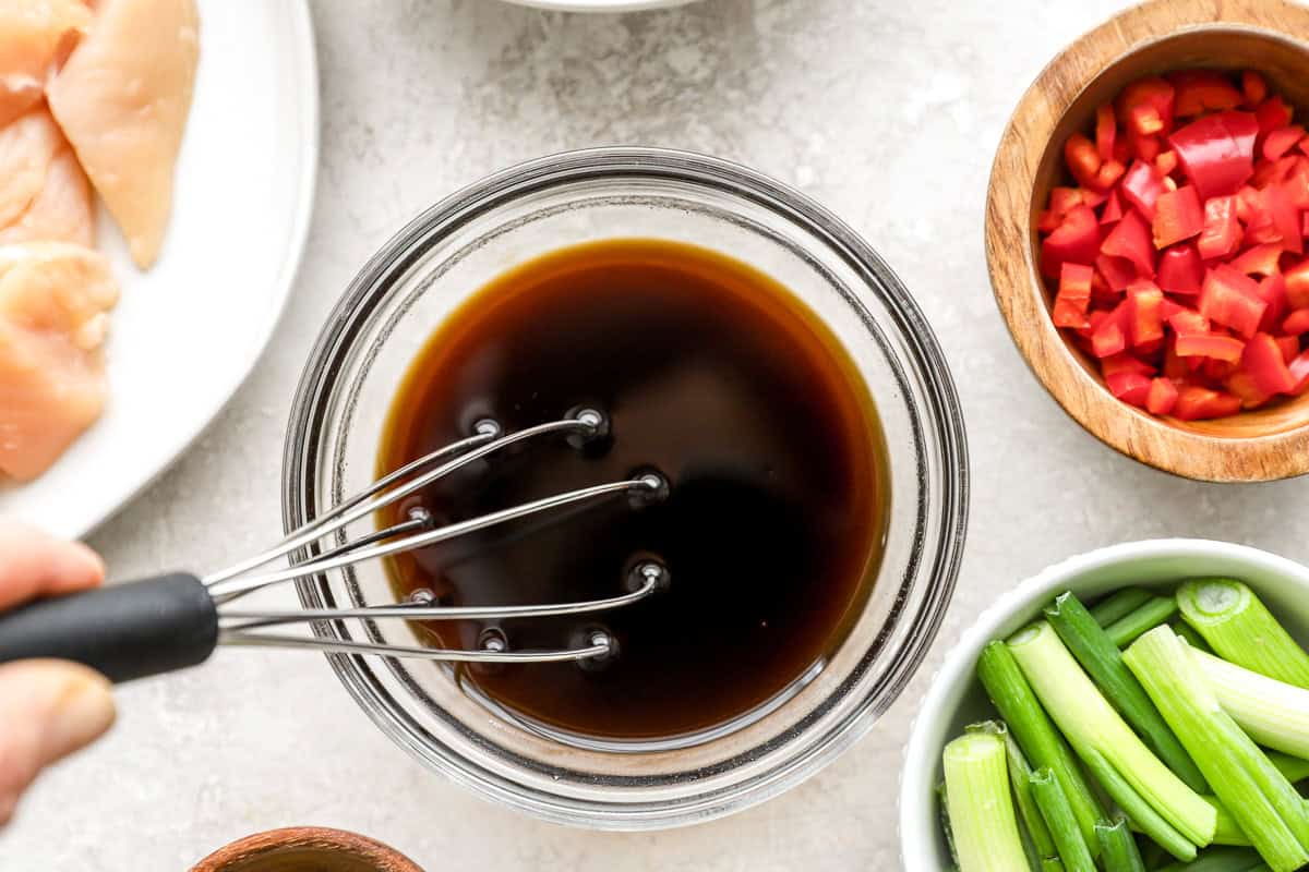 Glass bowl of dark stir-fry sauce being whisked, surrounded by chopped red chilies, green onions, and raw chicken pieces.