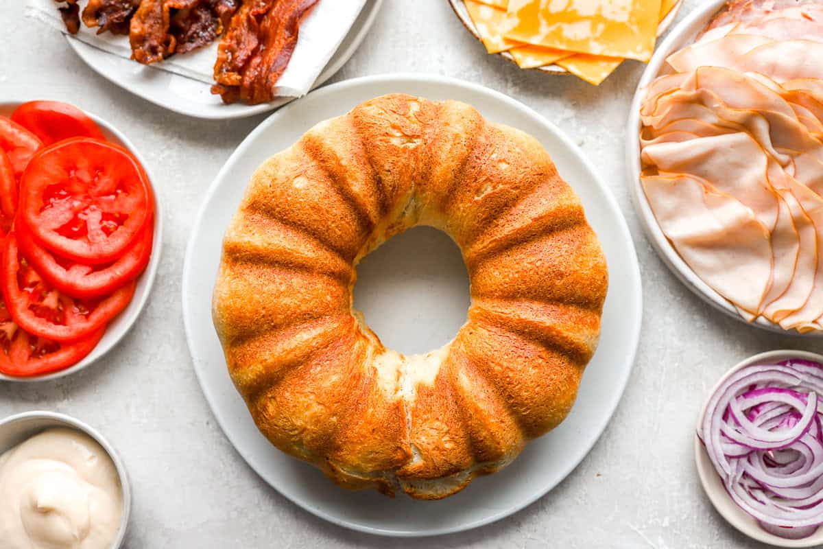 A golden-brown Bundt-shaped loaf of bread sits on a plate, ready to be sliced and turned into a giant party sandwich. Fresh sandwich ingredients surround it.