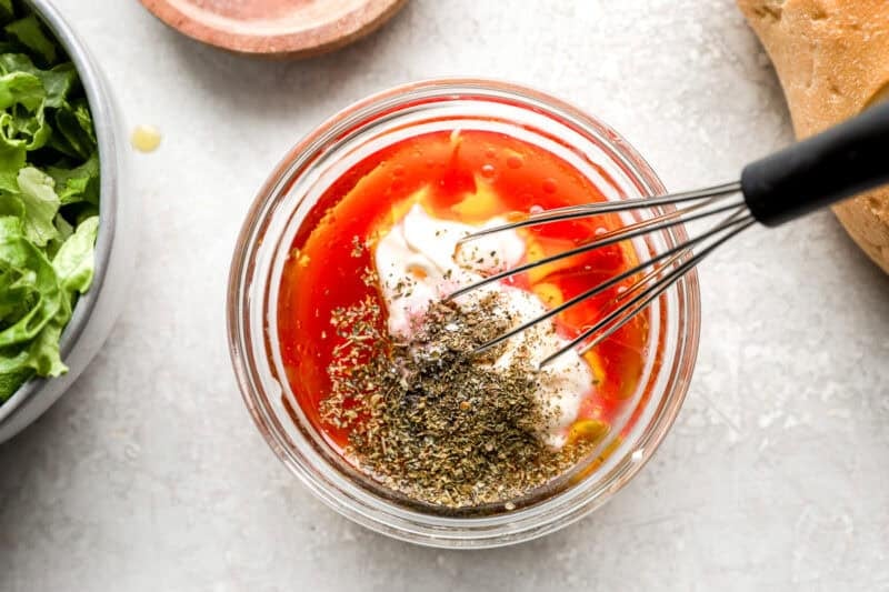 A small glass jar with mayonnaise, red wine vinegar, olive oil, and dried herbs being whisked together to create a creamy Italian dressing.