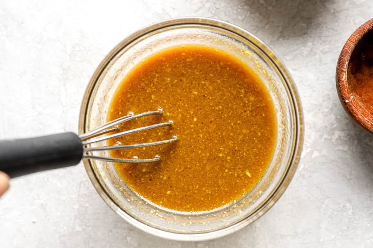 A close-up of a whisk mixing the maple Dijon glaze in a glass bowl, showing its rich golden-brown color and slightly thick consistency.
