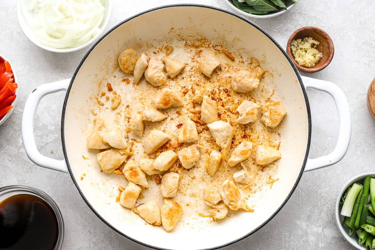 Chunks of browned chicken cooking in a white enamel skillet, surrounded by prepped vegetables and sauce ingredients.