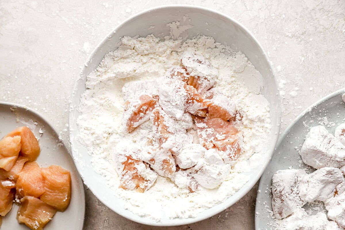 Raw chicken pieces being coated in cornstarch mixture in a bowl, with more chicken pieces on a plate nearby.