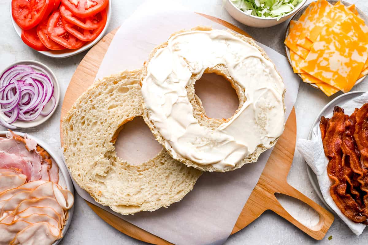 The Bundt-shaped bread has been sliced in half, with the inside spread generously with mayonnaise, sitting on a wooden cutting board with sandwich ingredients nearby.