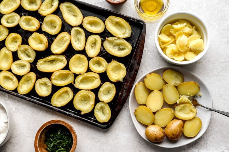 Sliced baby potatoes with the centers scooped out, laid out on a baking tray next to bowls of potato flesh, sour cream, and green onions.