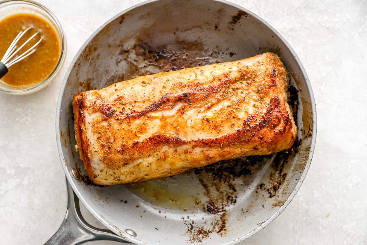 A pork loin being seared in a stainless steel skillet, developing a crispy golden-brown crust on the surface. A small bowl of maple Dijon glaze sits beside it.