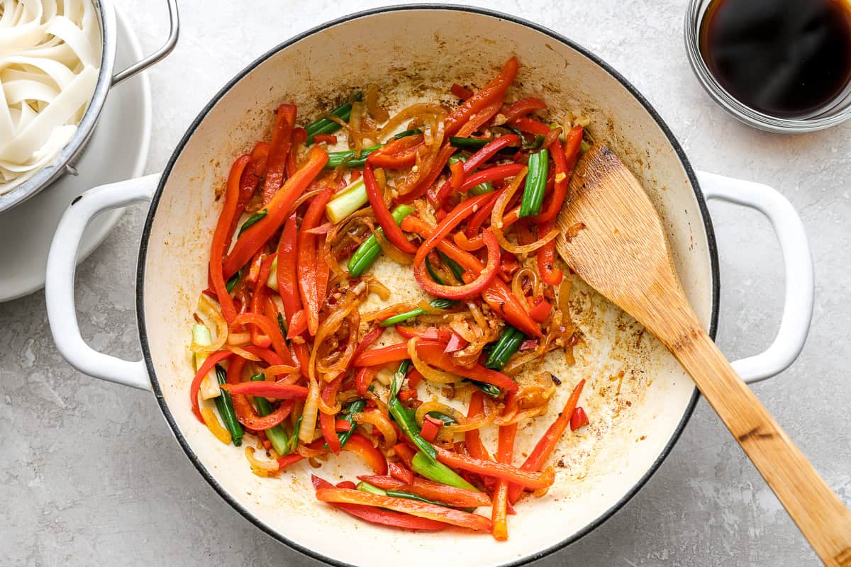 Red bell peppers, onions, and green onions being stir-fried in a skillet with visible caramelization on the edges.