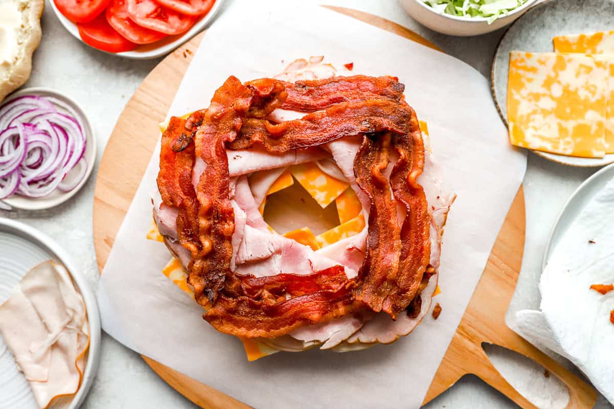 The sandwich assembly begins with a layer of Colby Jack cheese, turkey, ham, and crispy bacon stacked on the bottom half of the Bundt-shaped bread.