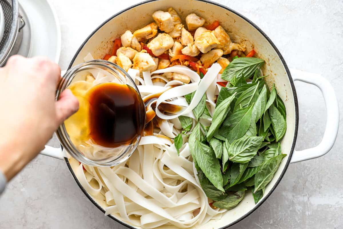 Sauce being poured over rice noodles, chicken, and fresh Thai basil in a large skillet during the final step of cooking.