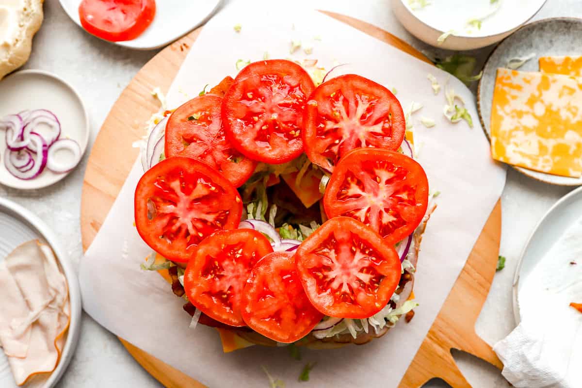 Fresh tomato slices are added on top of the stacked sandwich ingredients, along with thinly sliced red onions and shredded lettuce.