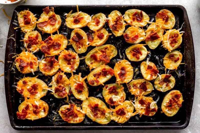 Potato skins filled with shredded cheddar cheese and bacon bits before the final bake, lined up on a sheet pan.