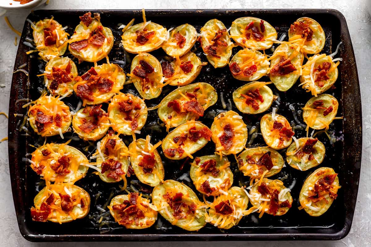 Potato skins filled with shredded cheddar cheese and bacon bits before the final bake, lined up on a sheet pan.