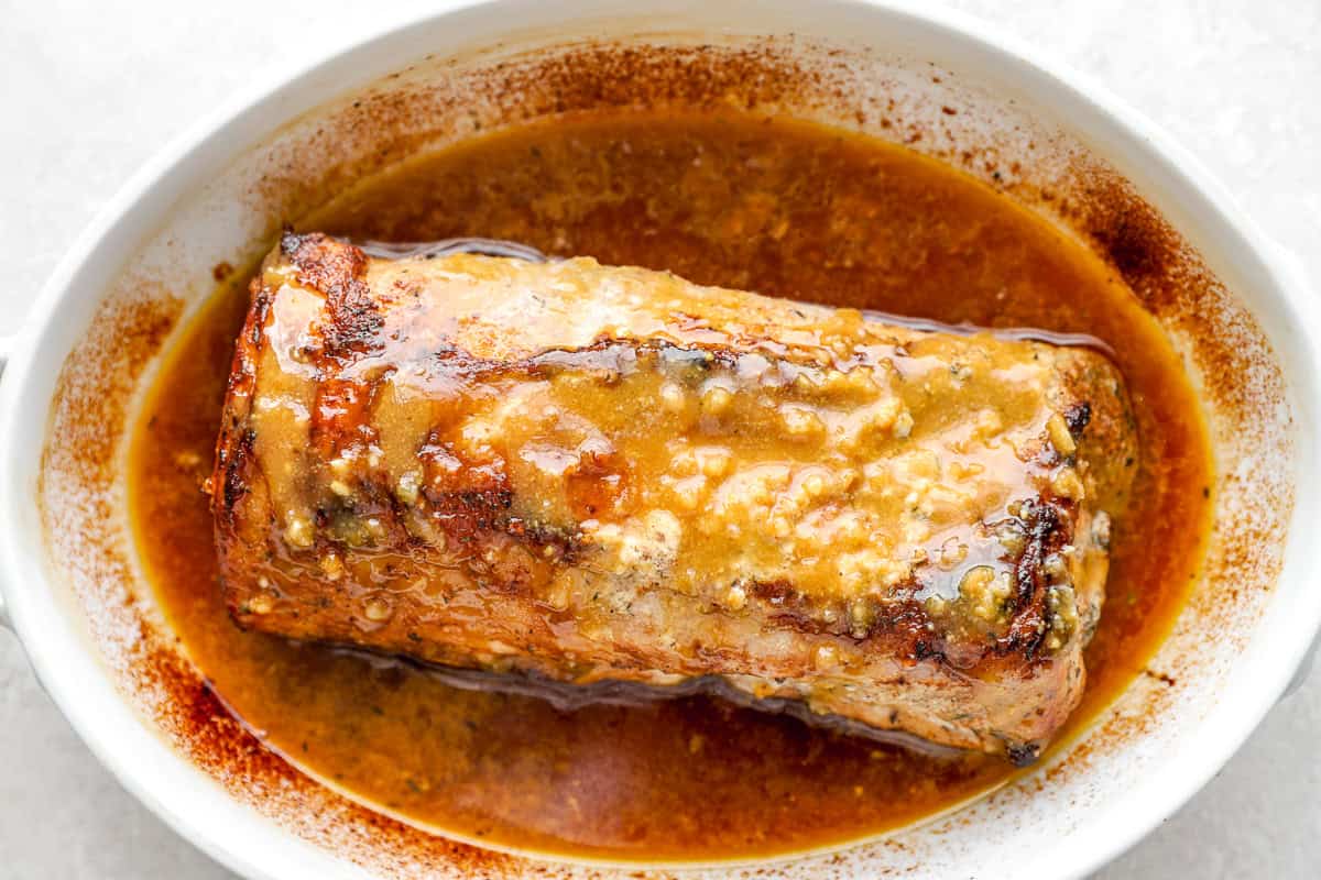 The pork loin in the baking dish after roasting, covered in a thick, caramelized maple Dijon sauce with visible minced garlic.