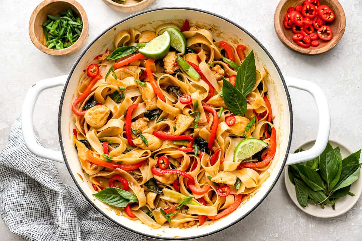 Finished drunken noodles in a large white skillet, garnished with fresh basil leaves, lime wedges, and sliced chilies.