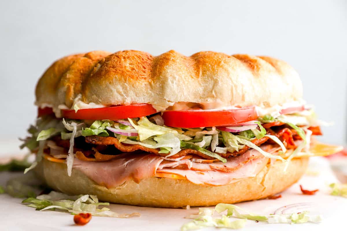 The fully assembled giant party sandwich is placed on a cutting board, showing off its stacked layers of deli meats, cheese, crispy bacon, lettuce, and tomato between a golden-brown Bundt-style loaf.