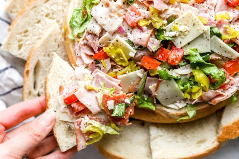 A hand lifting a piece of Italian bread loaded with the creamy, colorful hoagie dip, showcasing the textures of the meats, cheese, and vegetables.