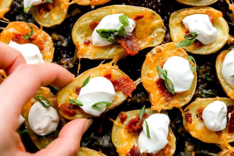 Close-up of a hand picking up one loaded potato skin bite with cheese, bacon, sour cream, and green onion.
