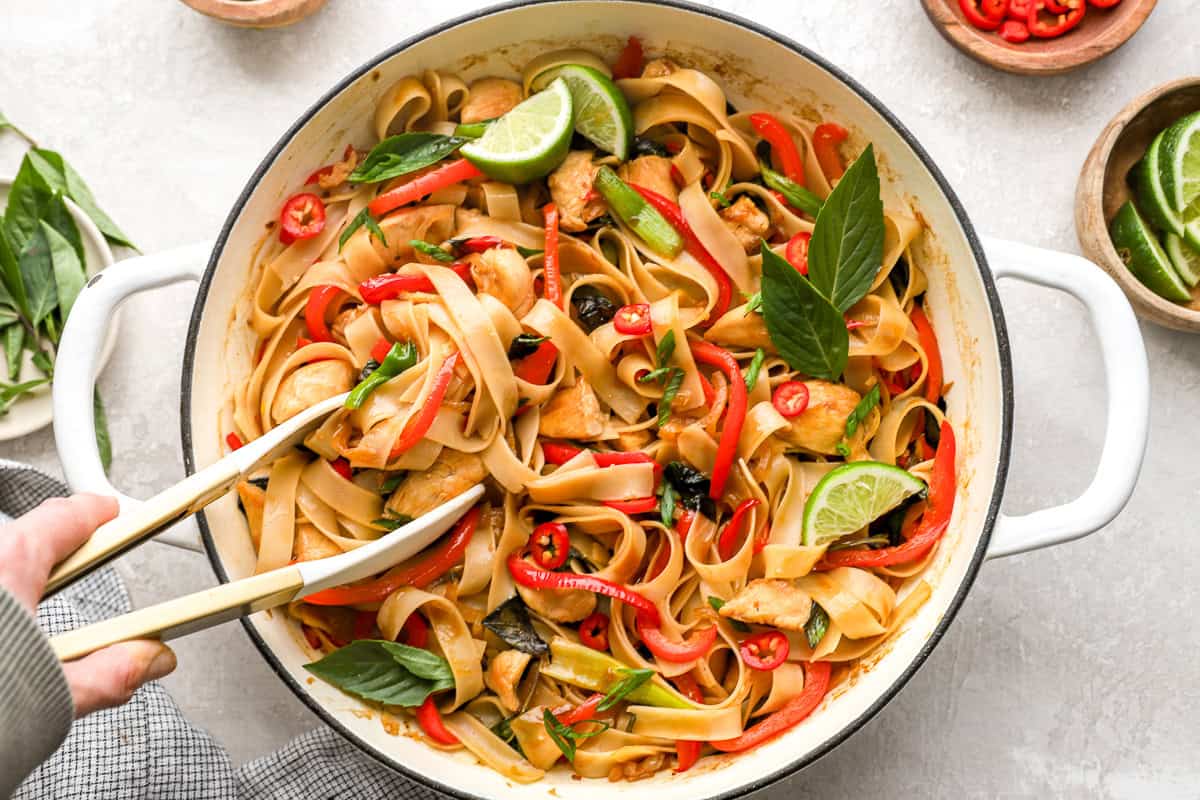Hand using tongs to toss cooked drunken noodles with chicken, Thai basil, and vegetables in a skillet.