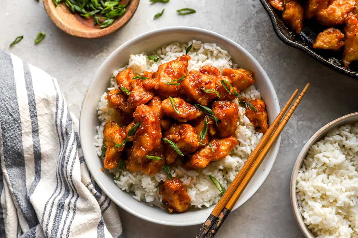 General Tso’s Chicken served in a bowl over rice with chopsticks and extra bowls of rice and scallions nearby.