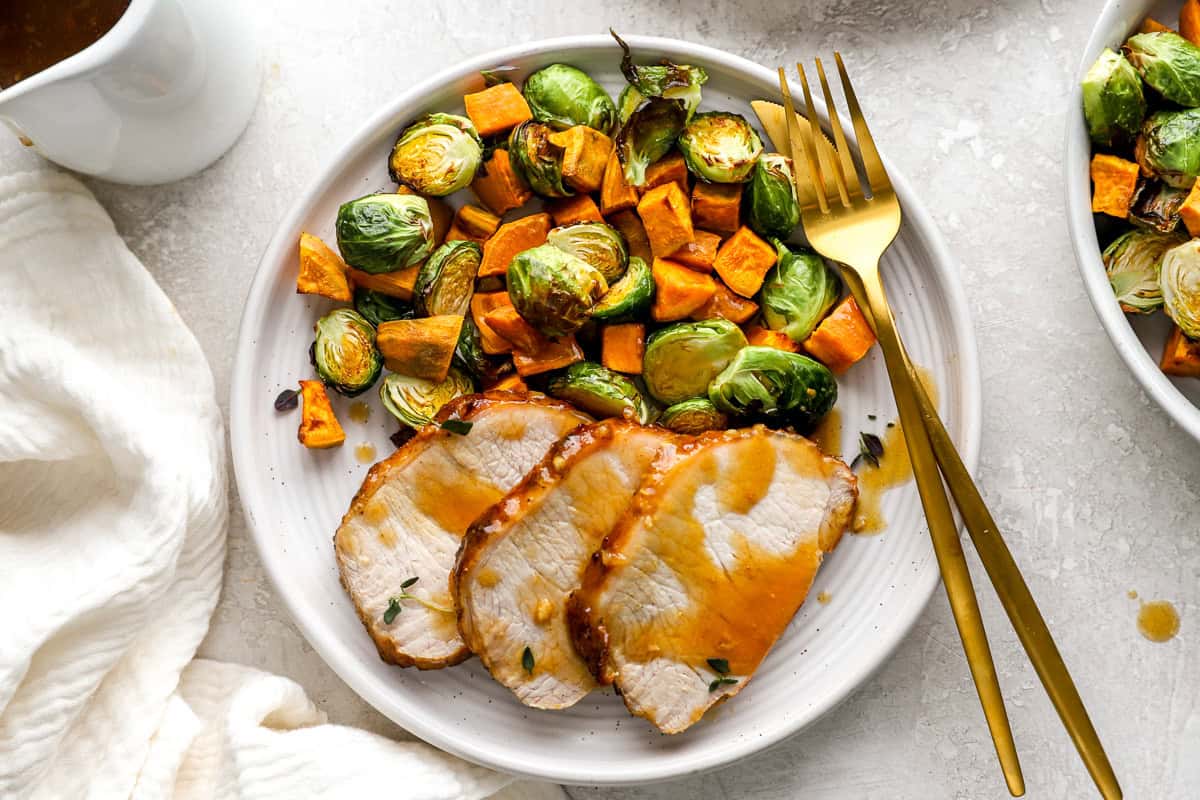 A plated meal featuring sliced pork loin with maple Dijon glaze, served alongside roasted Brussels sprouts and sweet potatoes, with gold utensils resting on the plate.