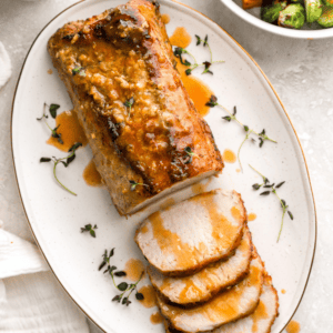 A beautifully roasted pork loin on a white oval platter, sliced and drizzled with a golden maple Dijon glaze, garnished with fresh thyme.