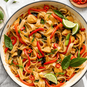 Close-up overhead shot of drunken noodles in a white skillet, featuring wide rice noodles, chicken, red bell peppers, Thai basil, sliced red chili, and lime wedges.