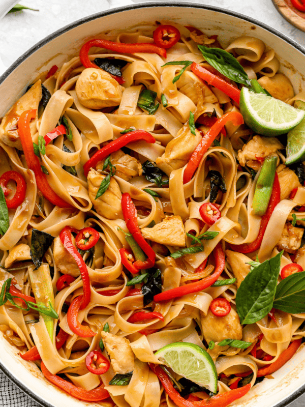 Close-up overhead shot of drunken noodles in a white skillet, featuring wide rice noodles, chicken, red bell peppers, Thai basil, sliced red chili, and lime wedges.