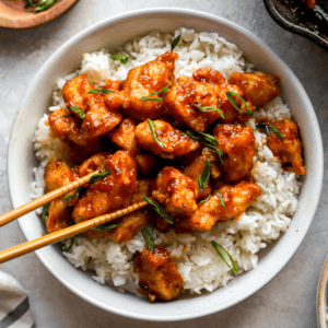 Bowl of crispy General Tso’s Chicken served over white rice and garnished with sliced green onions, with chopsticks resting on top.